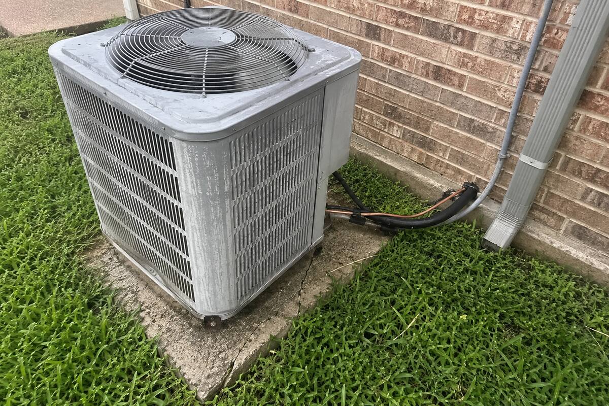 Weathered outdoor condenser unit sitting on a cracked concrete pad beside a brick wall